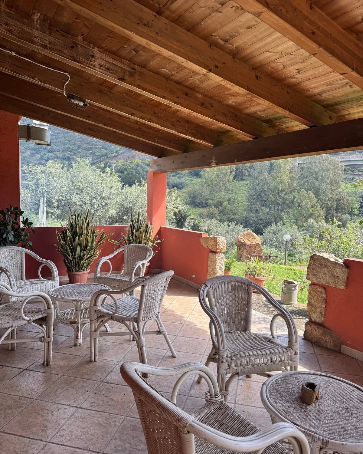 Veranda dell'Agriturismo Gutturus con travi in legno, poltroncine bianche in vimini, vista su ulivi e colline